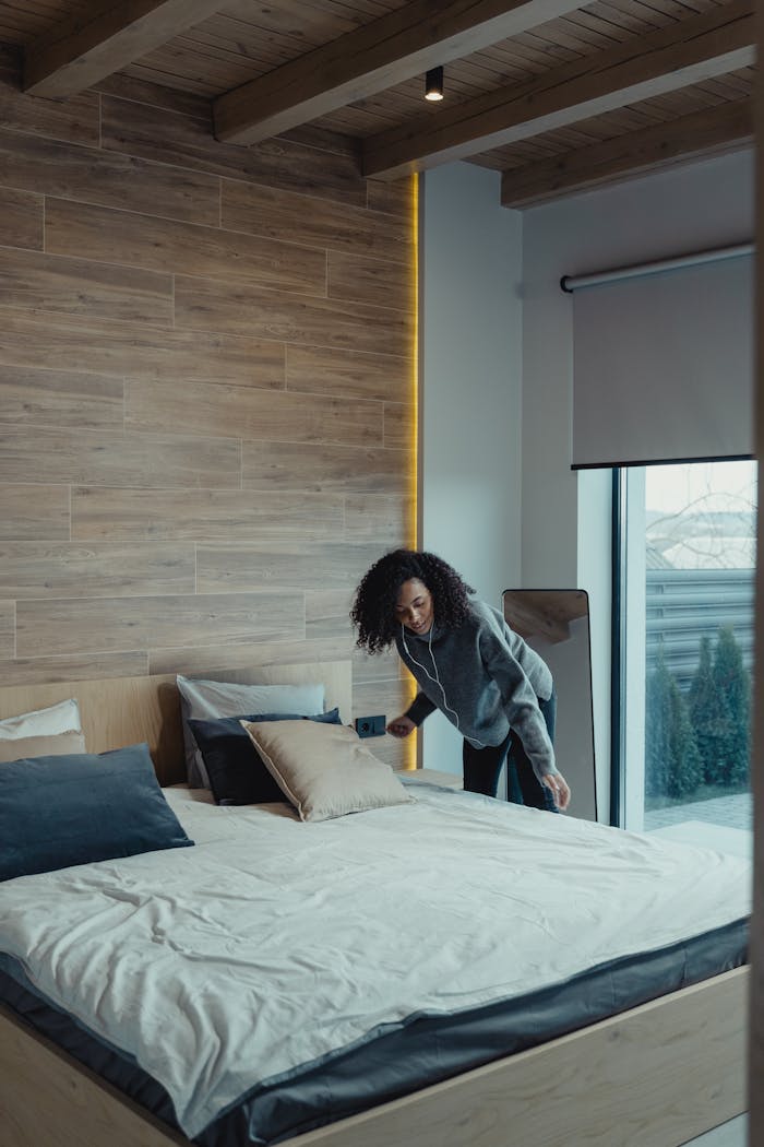 about-01 African American woman fixing pillows in a modern bedroom with wooden accents, promoting cleanliness and organization.